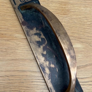 May include: An antique bronze door pull with the word "PULL" embossed on the top. The handle is curved and has a dark patina, showing signs of age. The rectangular plate has screw holes for mounting. A vintage hardware piece.