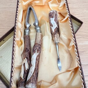 May include: A vintage nutcracker set comprising a nutcracker, a pick, and a small knife. The handles feature a white relief design on a brown background. The set is presented in a brown box with a gold satin lining.