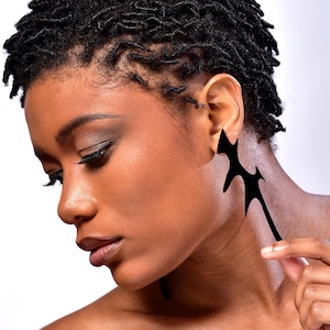 May include: A woman with short, tightly curled black hair is wearing a large, black, asymmetrical earring. The earring is shaped like a jagged, abstract design.