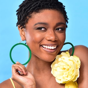 May include: A woman with short, dark hair wearing large green hoop earrings and a yellow top with a large yellow flower pinned to it. She is smiling and looking to the side.