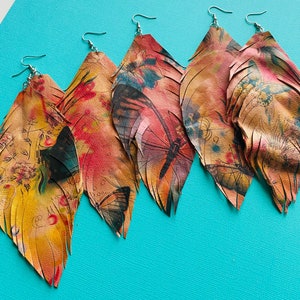 May include: Four colorful leather earrings with a fringed design. Each earring features a different butterfly and dragonfly pattern with shades of pink, yellow, orange, and brown.