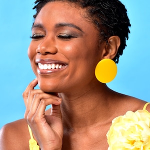 May include: A woman with short, dark hair is smiling and wearing large, bright yellow circular earrings. She is wearing a yellow top with a yellow flower on the shoulder.