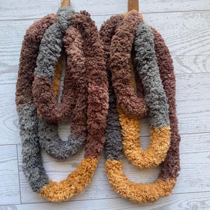 May include: A pair of handmade, oversized earrings. The earrings are made of a thick, looped yarn in shades of brown, gray, and gold. The earrings are suspended from silver-colored hooks and brown leather-like material.