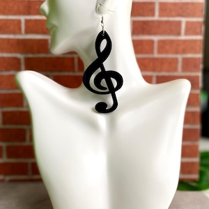 May include: Black treble clef earrings hanging from silver hooks. The earrings are displayed on a white mannequin against a red brick background. The musical note design is a simple, elegant shape.