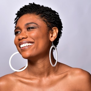 Könnte beinhalten: Eine Frau mit kurzen, dunklen Haaren lächelt und trägt große weiße Creolen.