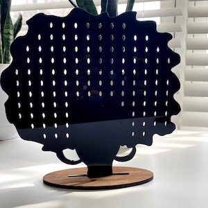 May include: Black earring holder shaped like a silhouette of a person with an afro hairstyle. The holder has multiple holes for earrings and is mounted on a wooden base.