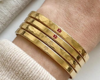 May include: A set of four gold-toned metal cuff bracelets with a hammered texture. Two of the bracelets feature a small red medical alert symbol, while the other two display a small engraved star-like icon on the outer surface of the metal.