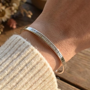 Op de afbeelding: Een zilveren manchetarmband met de gegraveerde tekst "FOR HER I WOULD RISK IT ALL". De armband wordt om een pols gedragen, met een beige mouw van een trui zichtbaar. Een klein boeket droogbloemen en een keramische mok staan op de achtergrond.