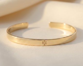 May include: A gold-toned metal cuff bracelet featuring a smooth, polished surface and an open-ended design. The center of the band is engraved with a small, six-pointed Star of Life medical symbol, indicating a medical alert identification piece.