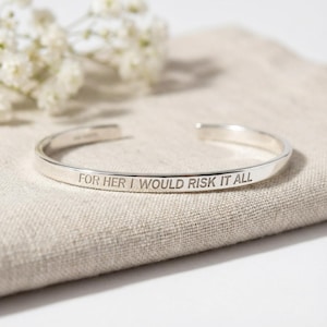 Può includere: Un bracciale rigido in metallo color argento dal design aperto. La superficie esterna del bracciale è incisa con il testo FOR HER I WOULD RISK IT ALL in un carattere pulito e senza grazie. Il bracciale ha una finitura liscia e lucida.