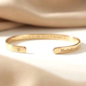 Puede incluir: Una pulsera de brazalete abierta de color dorado. La pulsera tiene una huella de pata en un extremo y la palabra "Meatball" grabada en el otro. La pulsera también tiene la inscripción "Forever by my side, forever in my heart."