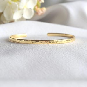 Peut inclure: Un bracelet manchette doré avec une texture martelée. Le bracelet ouvert est présenté sur une surface blanche, avec un arrière-plan doux et flou. Ce bijou est simple et élégant.