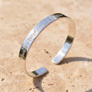 Op de afbeelding: Een zilveren manchetarmband met de inscriptie "When two become one. 9-26-2020." De armband heeft een gehamerde textuur en een open ontwerp. De armband wordt getoond op een lichtbruin oppervlak.