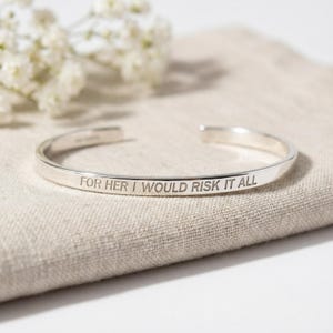 May include: A silver cuff bracelet with the engraved inscription "FOR HER I WOULD RISK IT ALL." The bracelet is displayed on a beige linen surface, with white flowers in the background.