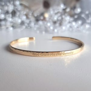 Può includere: Un bracciale a polsino aperto color oro con il testo inciso "God Grant Me The Serenity". Il bracciale ha una texture martellata ed è esposto su una superficie bianca. È un gioiello.