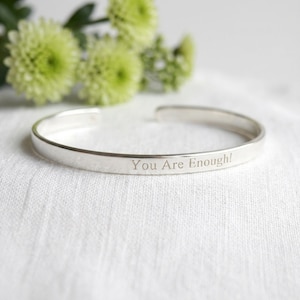 May include: A silver cuff bracelet with the engraved text "You Are Enough!" is displayed on a white surface. The open-ended bracelet is in focus, with a blurred background of green flowers.