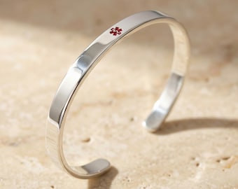 May include: A polished silver cuff bracelet with a red medical alert symbol. The bracelet has a smooth, rounded design and is shown on a neutral-toned surface. The medical symbol is a red cross-like emblem.