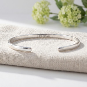 Op de afbeelding: Een zilverkleurige manchetarmband met de gegraveerde tekst "I am enough". De open armband wordt gepresenteerd op een beige linnen oppervlak, met zachte groene bloemen op de achtergrond. De armband heeft een eenvoudig, elegant ontwerp.