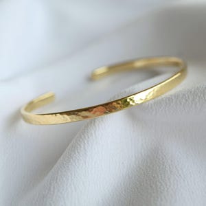 Könnte beinhalten: Ein goldfarbenes Armreif-Armband mit gehämmertem Muster. Das offene Armband wird auf einer weißen, strukturierten Oberfläche gezeigt. Das Schmuckstück hat ein schlichtes, elegantes Design.