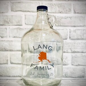 K&ouml;nnte beinhalten: Ein klares Glasgef&auml;&szlig; mit einem blauen Deckel und einem Griff. Das Gef&auml;&szlig; tr&auml;gt ein Etikett mit der Aufschrift "Lang Family Est. January 25" und eine orangefarbene Umrandung des Bundesstaates Alaska.