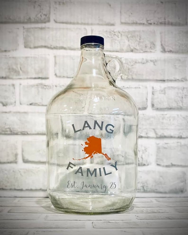 K&ouml;nnte beinhalten: Ein klares Glasgef&auml;&szlig; mit einem blauen Deckel und einem Griff. Das Gef&auml;&szlig; tr&auml;gt ein Etikett mit der Aufschrift "Lang Family Est. January 25" und eine orangefarbene Umrandung des Bundesstaates Alaska.