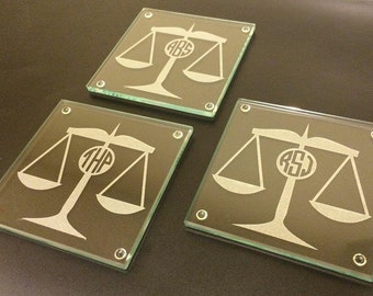 May include: Three glass coasters with a scale of justice design and personalized monograms. The coasters are square and have a frosted glass finish.
