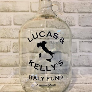 K&ouml;nnte beinhalten: Ein klares Glasgef&auml;&szlig; mit schwarzem Deckel und Henkel. Das Gef&auml;&szlig; hat einen schwarzen Aufkleber mit dem Text "Lucas & Kelly's Italy Fund" und einer Silhouette Italiens. Der Text "Adventure Awaits" befindet sich ebenfalls auf dem Aufkleber.