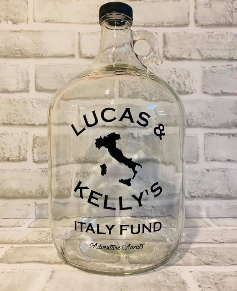 K&ouml;nnte beinhalten: Ein klares Glasgef&auml;&szlig; mit schwarzem Deckel und Henkel. Das Gef&auml;&szlig; hat einen schwarzen Aufkleber mit dem Text "Lucas & Kelly's Italy Fund" und einer Silhouette Italiens. Der Text "Adventure Awaits" befindet sich ebenfalls auf dem Aufkleber.
