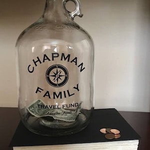 K&ouml;nnte beinhalten: Ein klares Glasgef&auml;&szlig; mit schwarzem Deckel und Henkel. Das Gef&auml;&szlig; hat ein schwarzes Etikett mit dem Text "Chapman Family Travel Fund Adventure Awaits". Auf dem Etikett befindet sich ein Kompass-Design und im Gef&auml;&szlig; befindet sich ein Dollar-Schein.