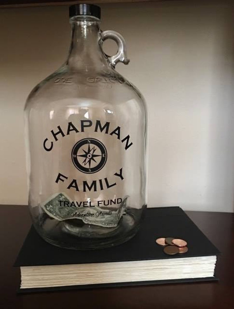 K&ouml;nnte beinhalten: Ein klares Glasgef&auml;&szlig; mit schwarzem Deckel und Henkel. Das Gef&auml;&szlig; hat ein schwarzes Etikett mit dem Text "Chapman Family Travel Fund Adventure Awaits". Auf dem Etikett befindet sich ein Kompass-Design und im Gef&auml;&szlig; befindet sich ein Dollar-Schein.