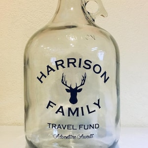 K&ouml;nnte beinhalten: Ein klares Glasgef&auml;&szlig; mit schwarzem Deckel und Henkel. Das Gef&auml;&szlig; hat einen schwarz-wei&szlig;en Aufkleber mit dem Text "HARRISON FAMILY TRAVEL FUND Adventure Awaits" und eine Silhouette eines Hirsches mit Geweih.