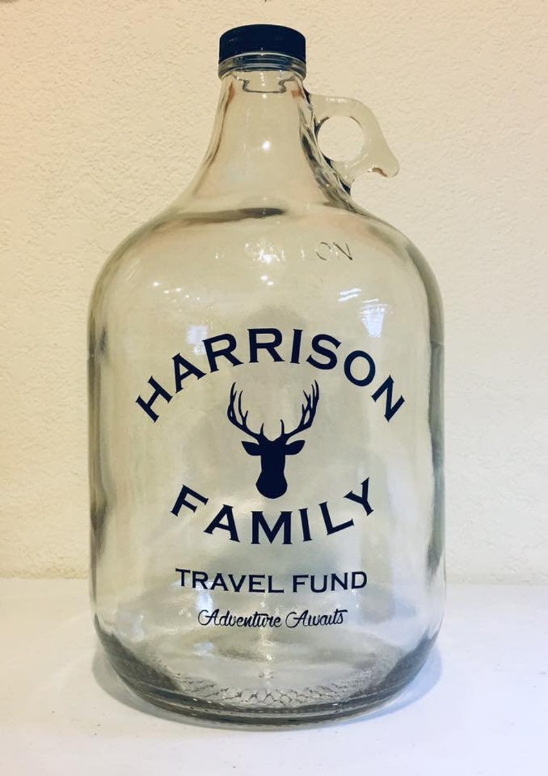 K&ouml;nnte beinhalten: Ein klares Glasgef&auml;&szlig; mit schwarzem Deckel und Henkel. Das Gef&auml;&szlig; hat einen schwarz-wei&szlig;en Aufkleber mit dem Text "HARRISON FAMILY TRAVEL FUND Adventure Awaits" und eine Silhouette eines Hirsches mit Geweih.