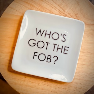 May include: A square white ceramic trinket dish with rounded corners featuring the black text WHO'S GOT THE FOB? printed in the center. The dish has a smooth, glossy finish and a simple, clean design suitable for holding small items like keys or jewelry.