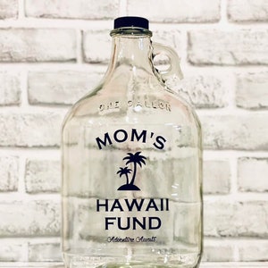 K&ouml;nnte beinhalten: Ein klares Glasgef&auml;&szlig; mit einem schwarzen Deckel und einem schwarzen Etikett mit der Aufschrift "Mamas Hawaii-Fonds Abenteuer wartet". Das Gef&auml;&szlig; hat einen Griff und steht auf einer wei&szlig;en Ziegelunterlage.