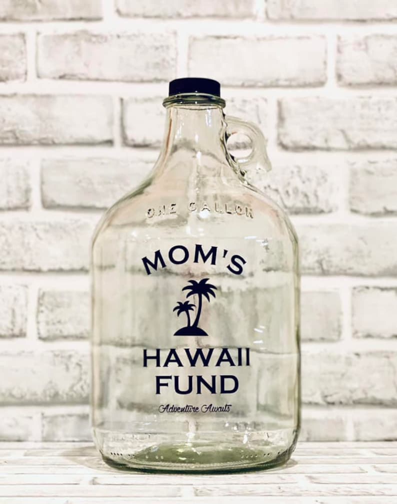K&ouml;nnte beinhalten: Ein klares Glasgef&auml;&szlig; mit einem schwarzen Deckel und einem schwarzen Etikett mit der Aufschrift "Mamas Hawaii-Fonds Abenteuer wartet". Das Gef&auml;&szlig; hat einen Griff und steht auf einer wei&szlig;en Ziegelunterlage.