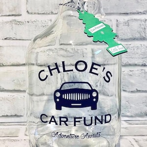 May include: Clear glass jug with a black lid, handle, and a green car air freshener tag. The jug has the words "CHLOE'S CAR FUND" and a car graphic, with "Adventure Awaits" below. The jug is on a white brick background.