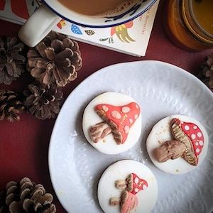 May include: Three white iced biscuits decorated with red and white fondant mushrooms. The biscuits are on a white plate with a decorative pattern.