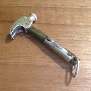 Op de afbeelding: Een kleine hamer in vintage-stijl met een zilveren kop en een messing en zilveren handvat. De hamer heeft een klauw om spijkers te verwijderen en een kleine ring aan het uiteinde van het handvat. Het ligt op een houten oppervlak.