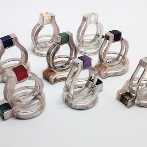 Puede incluir: Una colección de anillos de plata, cada uno con una piedra preciosa cuadrada de varios colores. Los anillos tienen un diseño moderno y geométrico con una base de doble banda y una sección superior curvada. Los colores de las piedras preciosas incluyen rojo, verde, azul y morado.