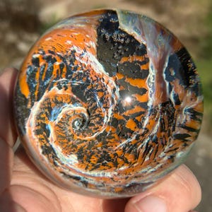 May include: A glass paperweight with a swirling pattern of black, orange, and white. The pattern resembles a spiral or vortex.