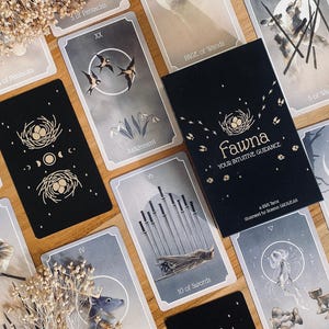 May include: A deck of Fawna tarot cards featuring black and grey cards with illustrations of birds, nests, and swords. The box is black with gold text reading Fawna Your Intuitive Guidance, a 78 card deck illustrated by Andrea Goulart, surrounded by dried flowers.