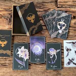 May include: A black box with gold accents and a guidebook for a tarot deck. The box has a bird design with a crescent moon and stars. The guidebook has the text "The Magpie's Lenormand Guidebook". There are five cards from the deck, including "The Lily", "The Moon", "The Anchor", and two cards with a bird design.