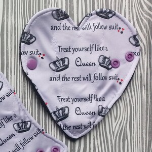 May include: Purple heart-shaped fabric with a white background and a repeating pattern of crowns and playing card suits. The text "Treat yourself like a Queen and the rest will follow suit" is printed in black on the fabric.
