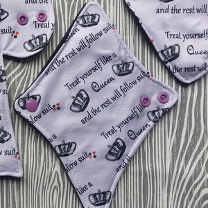 Puede incluir: Varios protectores menstruales de tela reutilizables de varias formas. Los protectores son blancos con un diseño repetido de corona y corazón, y el texto "Treat yourself like a Queen and the rest will follow suit." Cada protector tiene broches morados.