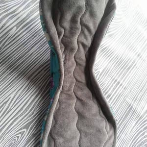May include: A gray and teal reusable cloth menstrual pad with a quilted gray absorbent layer and a floral print on the teal fabric.
