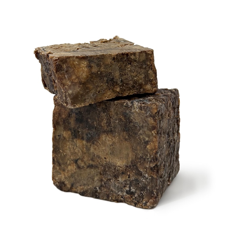 May include: Two bars of dark brown African black soap stacked on top of each other. The soap has a rough, textured surface.