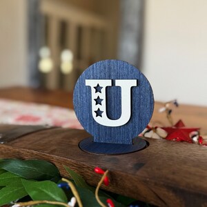 May include: A blue wooden circle with a white letter "U" and five white stars. The circle is on a wooden surface.