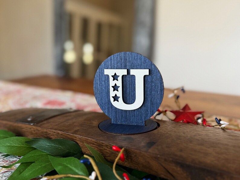 May include: A blue wooden circle with a white letter "U" and five white stars. The circle is on a wooden surface.