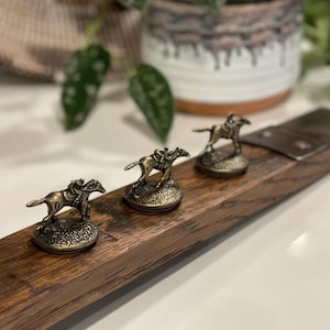 May include: A wooden board with four metal horse and jockey figurines attached. The board is dark brown with a natural wood grain. The figurines are gold in color and are arranged in a line.
