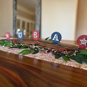 May include: A wooden centrepiece with five round candle holders, each decorated with a red, white and blue patriotic design. The holders spell out "USA" with a star on each end.